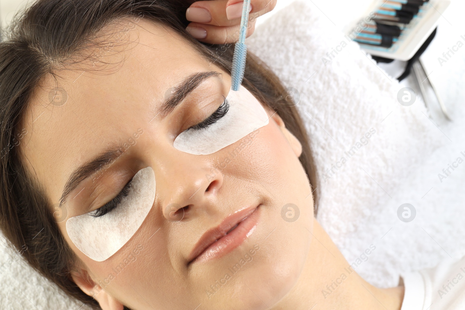 Photo of Esthetician brushing woman's lash extensions during procedure in beauty salon, closeup