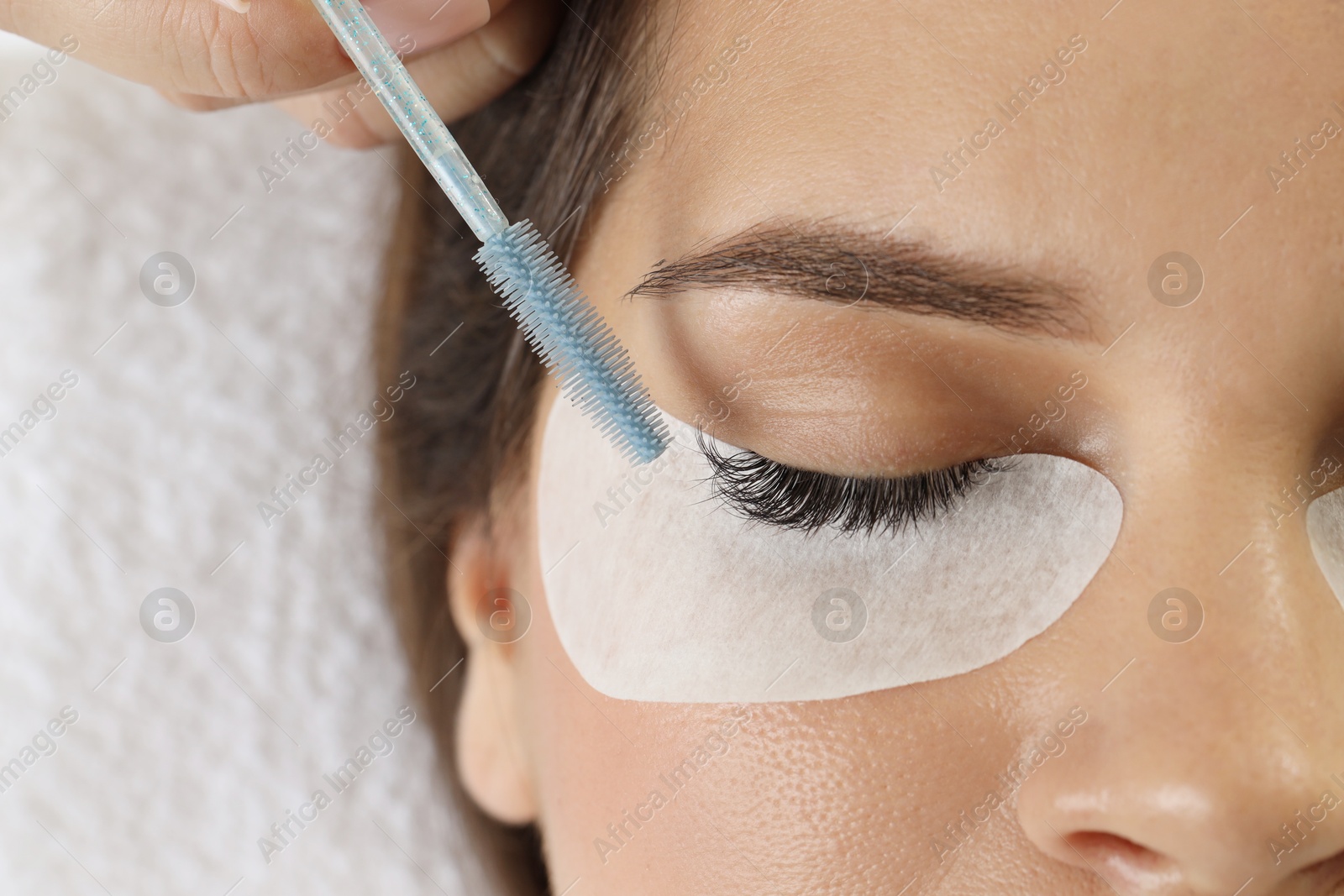 Photo of Esthetician brushing woman's lash extensions during procedure in beauty salon, closeup