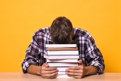 Young student with stack of books having stress before exam at wooden table against yellow background