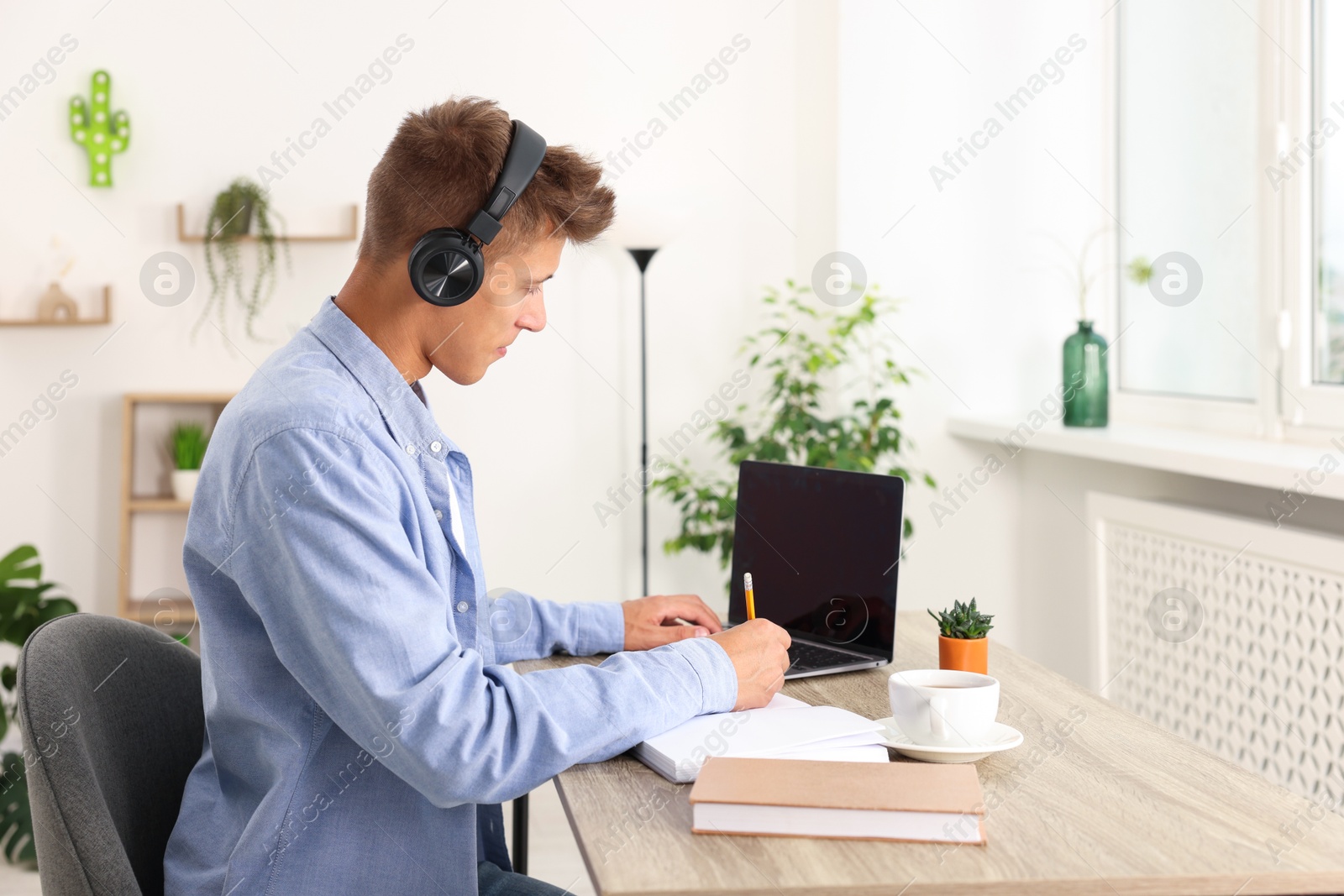 Photo of Student in headphones taking notes while studying with laptop at wooden table indoors