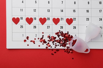 Calendar with marked dates and menstrual cups on red background, top view