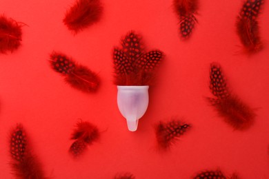 Photo of Menstrual cup with feathers on red background, flat lay