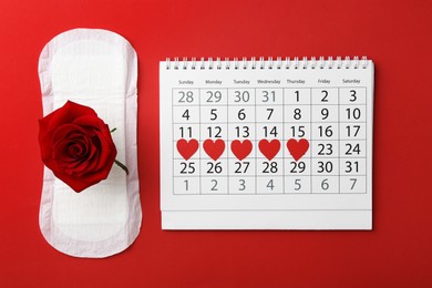 Photo of Calendar with marked menstrual cycle days, pad and rose on red background, flat lay