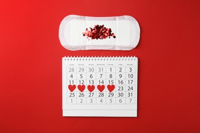 Calendar with marked menstrual cycle days and pad on red background, flat lay