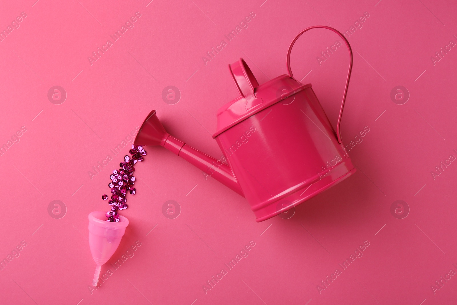 Photo of Pouring sequins into menstrual cup from watering can on pink background, top view