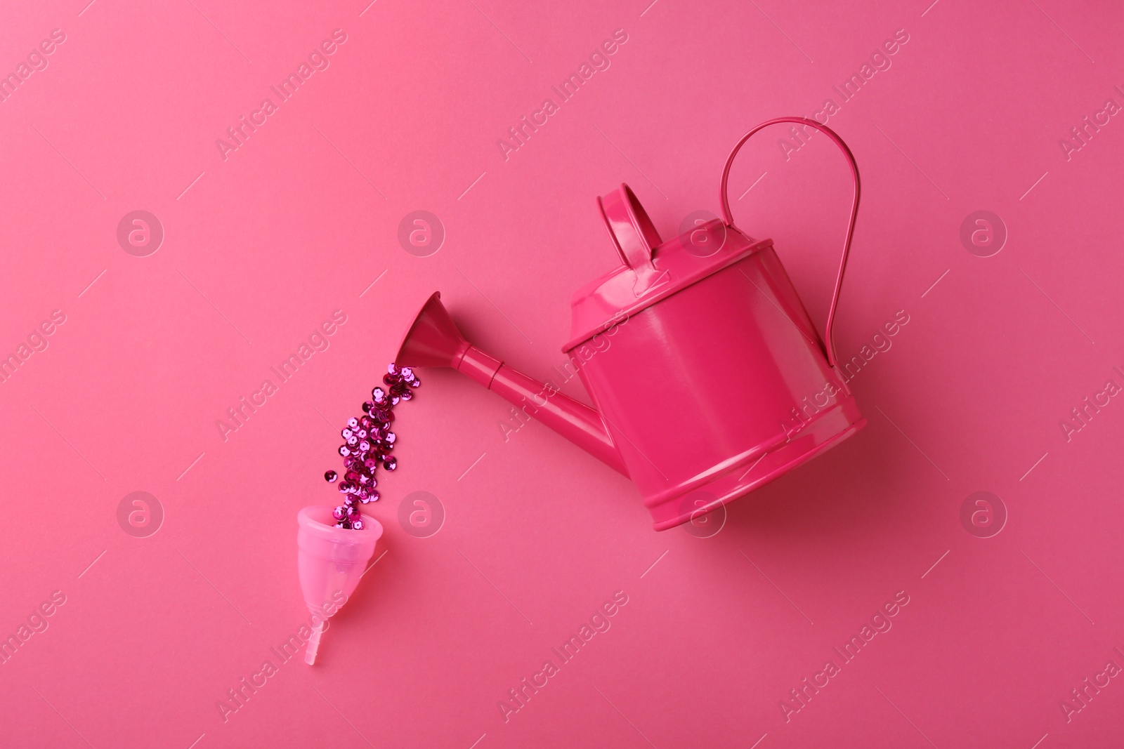 Photo of Pouring sequins into menstrual cup from watering can on pink background, top view