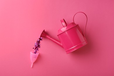 Photo of Pouring sequins into menstrual cup from watering can on pink background, top view