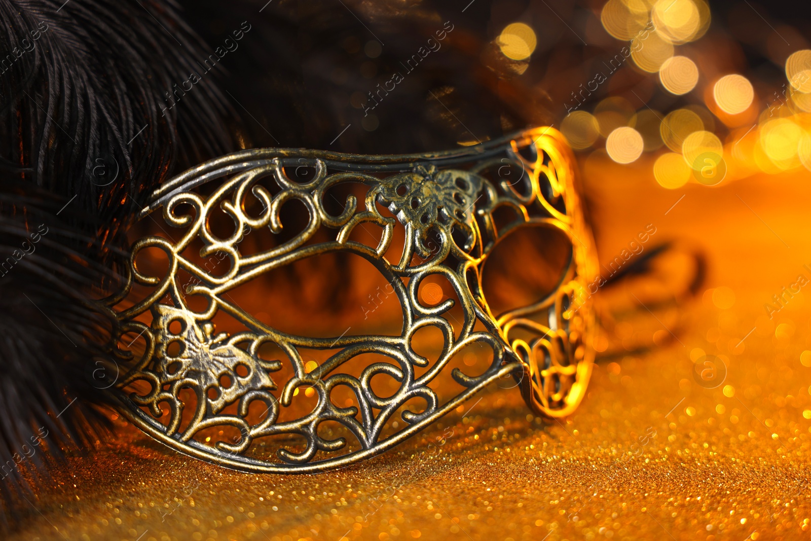 Photo of Beautiful carnival mask and black feathers on golden background against blurred lights, closeup. Bokeh effect