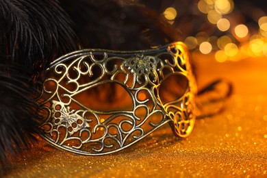 Photo of Beautiful carnival mask and black feathers on golden background against blurred lights, closeup. Bokeh effect