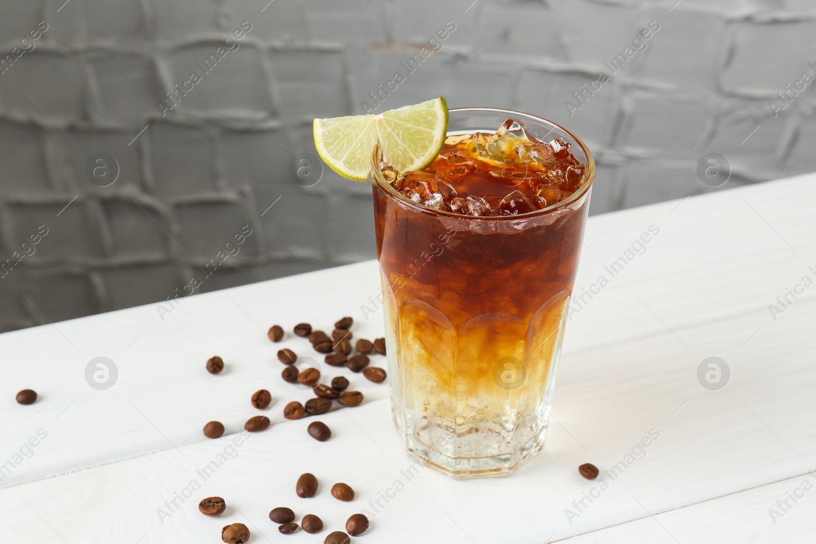 Photo of Refreshing espresso tonic drink with slice of lime and coffee beans on white wooden table