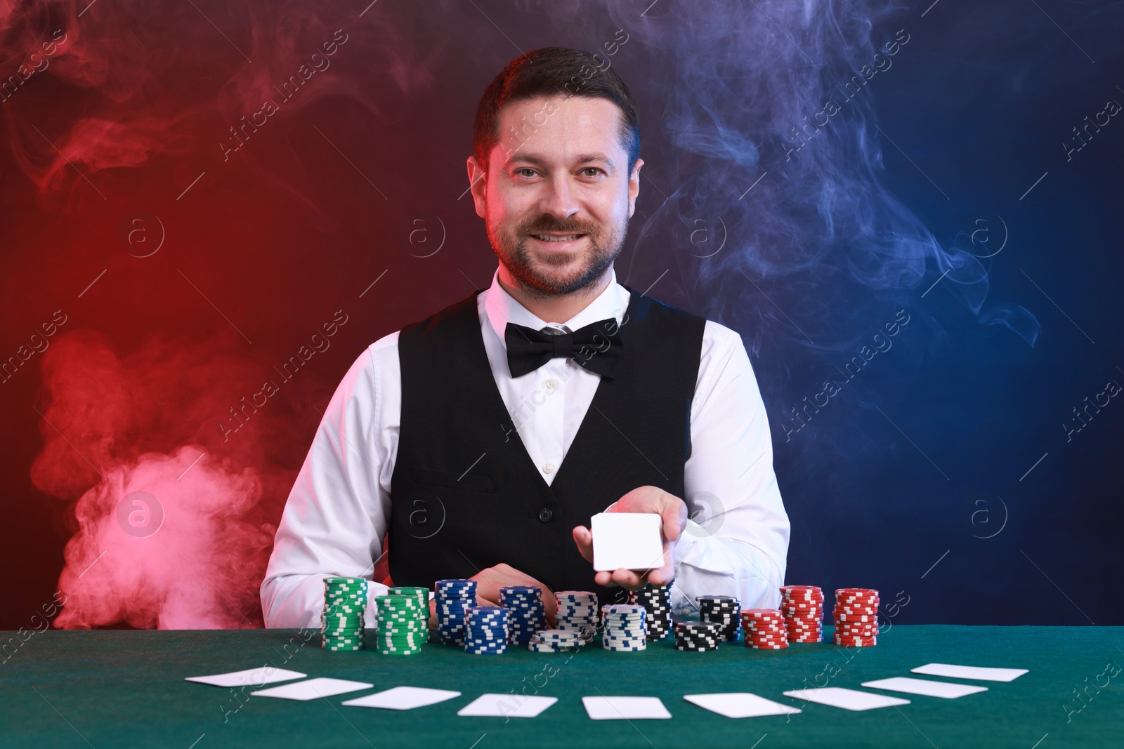 Photo of Professional croupier holding playing cards and casino chips at gambling table against color background with smoke