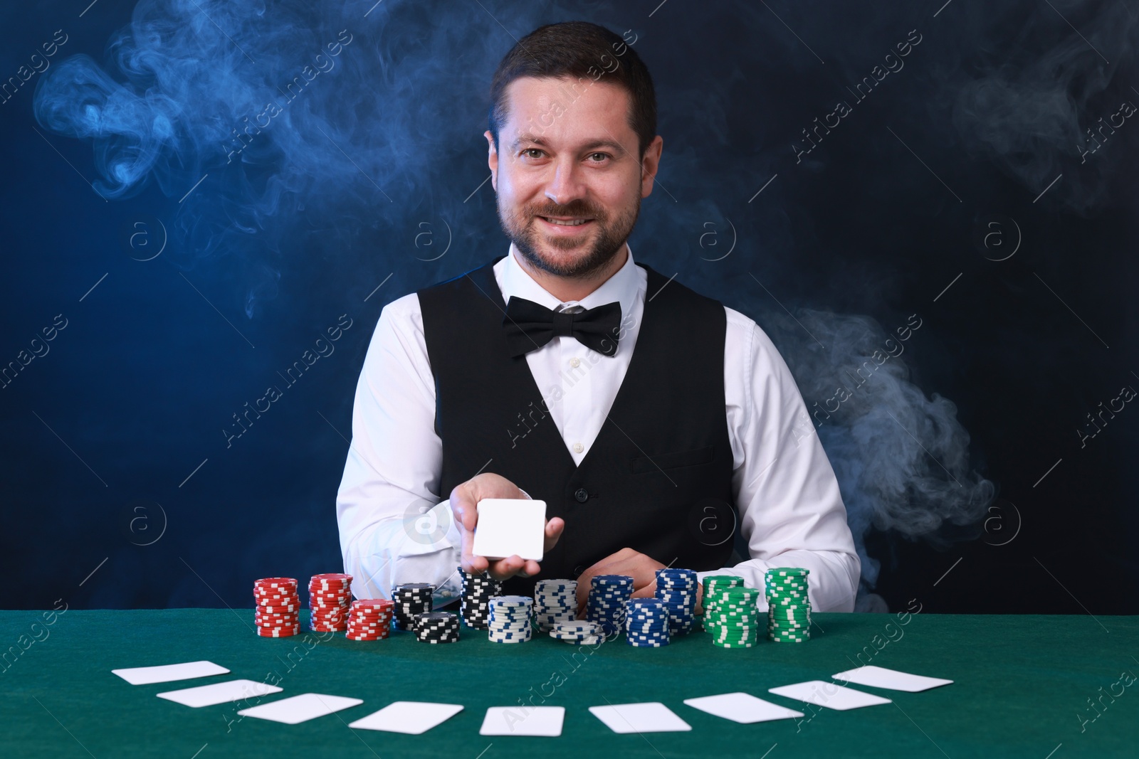 Photo of Professional croupier holding playing cards and casino chips at gambling table against color background with smoke