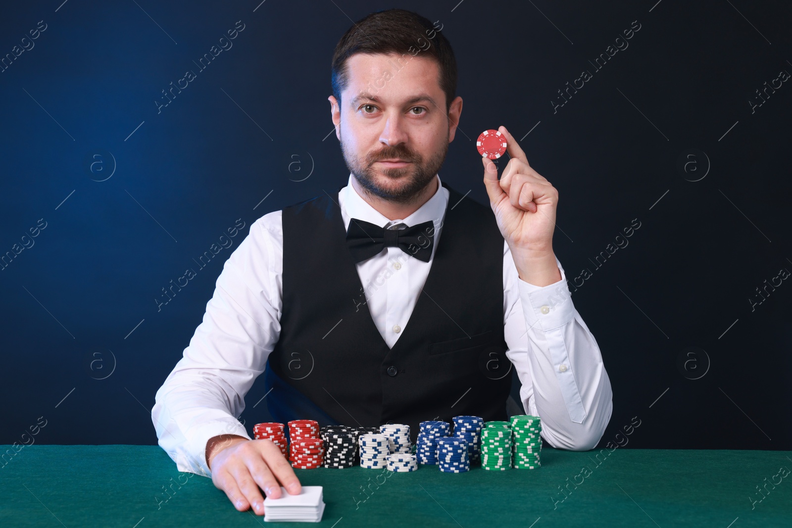 Photo of Professional croupier with playing cards and casino chips at gambling table against color background