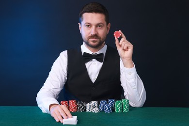 Professional croupier with playing cards and casino chips at gambling table against color background