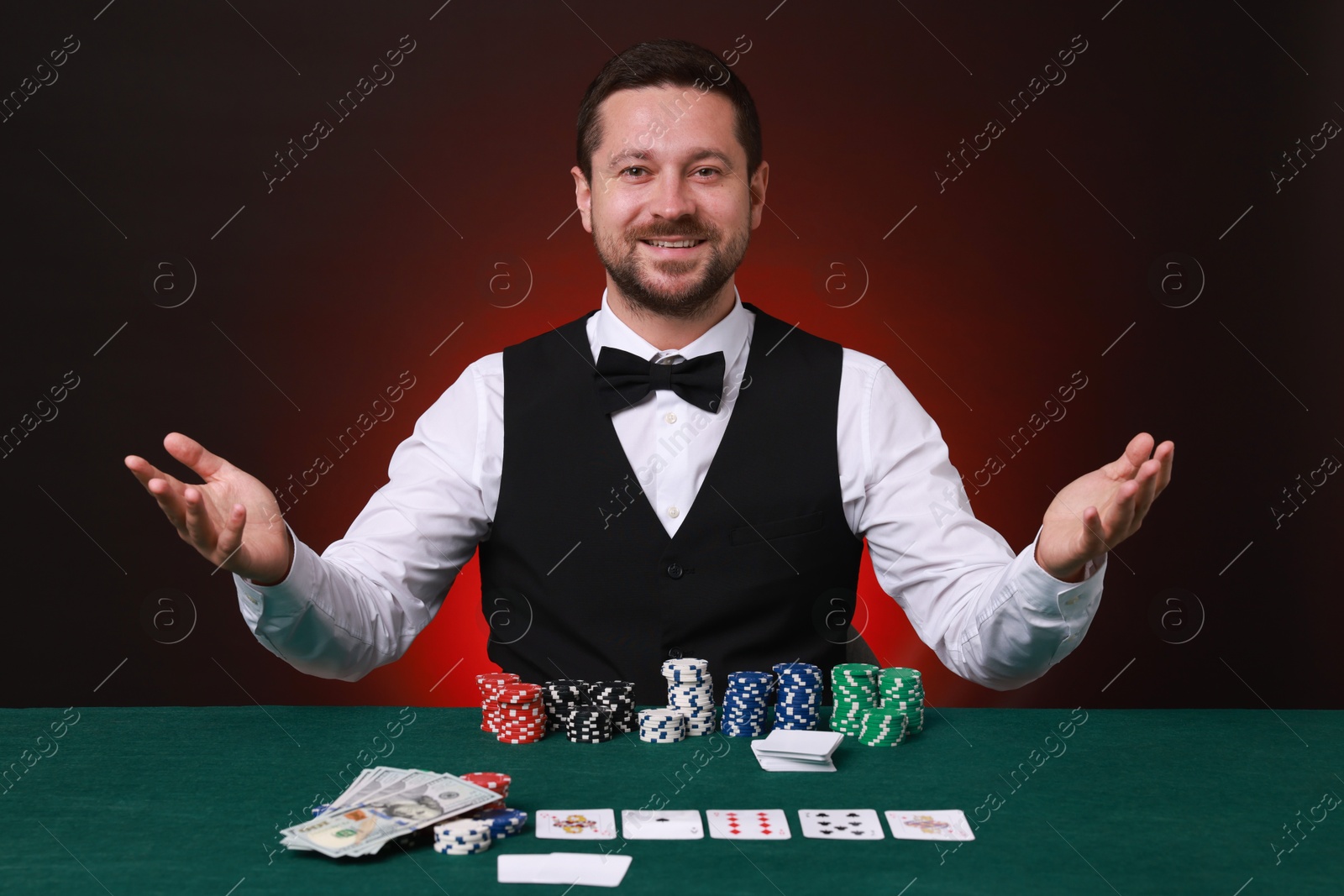 Photo of Professional croupier at gambling table with casino chips, playing cards and money against dark red background