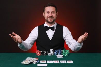 Photo of Professional croupier at gambling table with casino chips, playing cards and money against dark red background