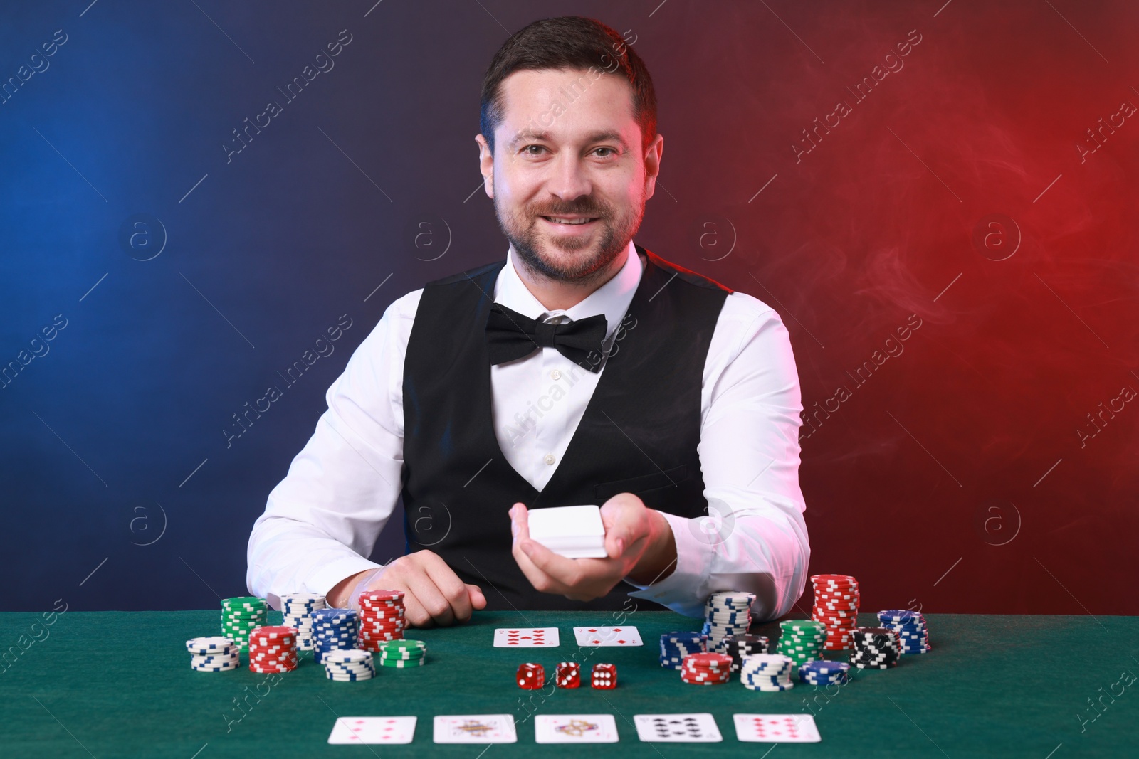 Photo of Professional croupier with playing cards at gambling table against color background with smoke