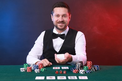 Photo of Professional croupier with playing cards at gambling table against color background with smoke