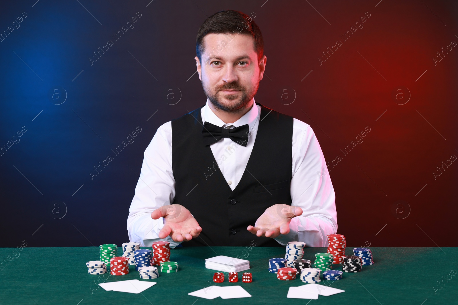 Photo of Professional croupier at gambling table with playing cards, casino chips and dice against color background