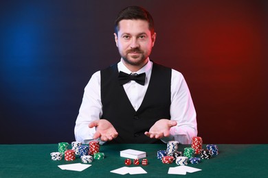 Photo of Professional croupier at gambling table with playing cards, casino chips and dice against color background