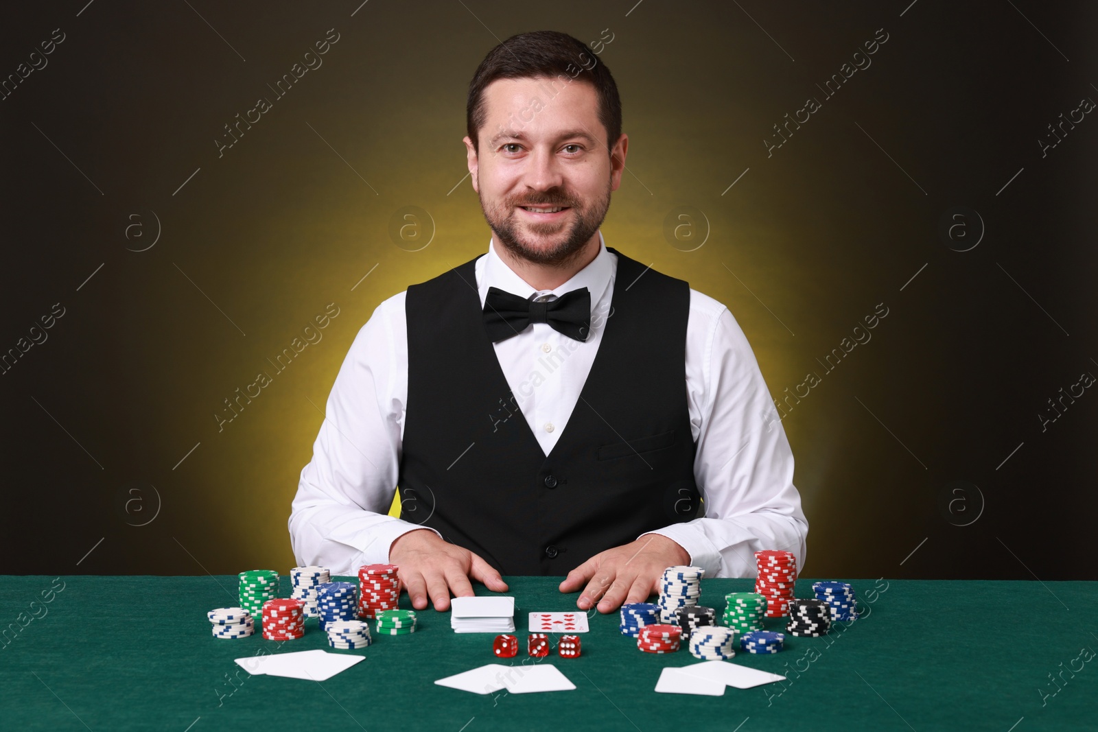 Photo of Professional croupier at gambling table with playing cards, casino chips and dice against dark yellow background