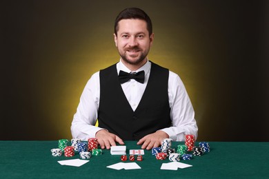 Photo of Professional croupier at gambling table with playing cards, casino chips and dice against dark yellow background