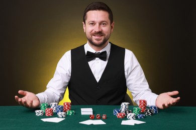 Professional croupier at gambling table with playing cards, casino chips and dice against dark yellow background