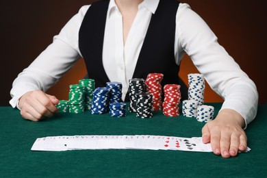 Photo of Professional croupier with casino chips and playing cards at gambling table on color background, closeup