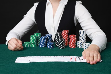 Photo of Professional croupier with casino chips and playing cards at gambling table on black background, closeup