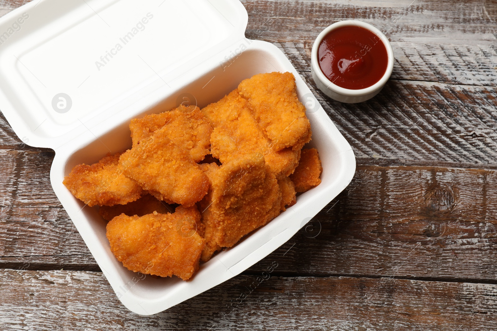 Photo of Delicious chicken nuggets in takeaway container and ketchup on wooden table, above view