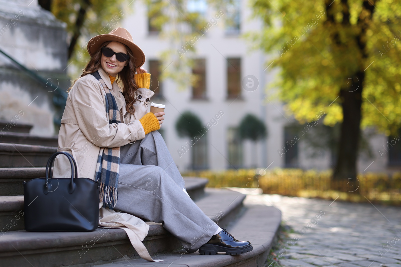 Photo of Charming young woman with cup of coffee on city street, space for text. Autumn season