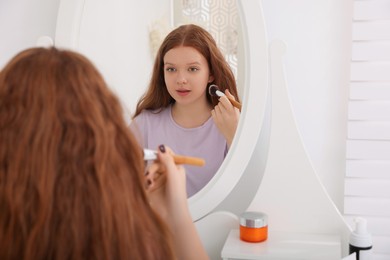 Photo of Teenage girl applying blush with brush near mirror indoors