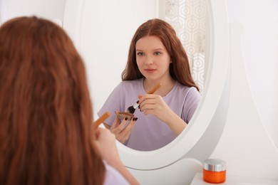 Teenage girl applying blush with brush near mirror indoors