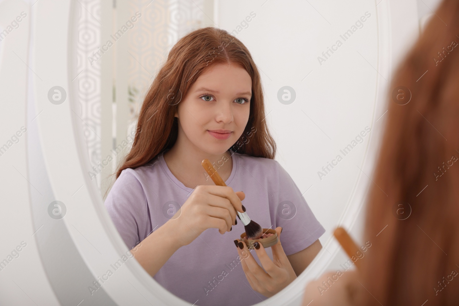 Photo of Teenage girl applying blush with brush near mirror indoors