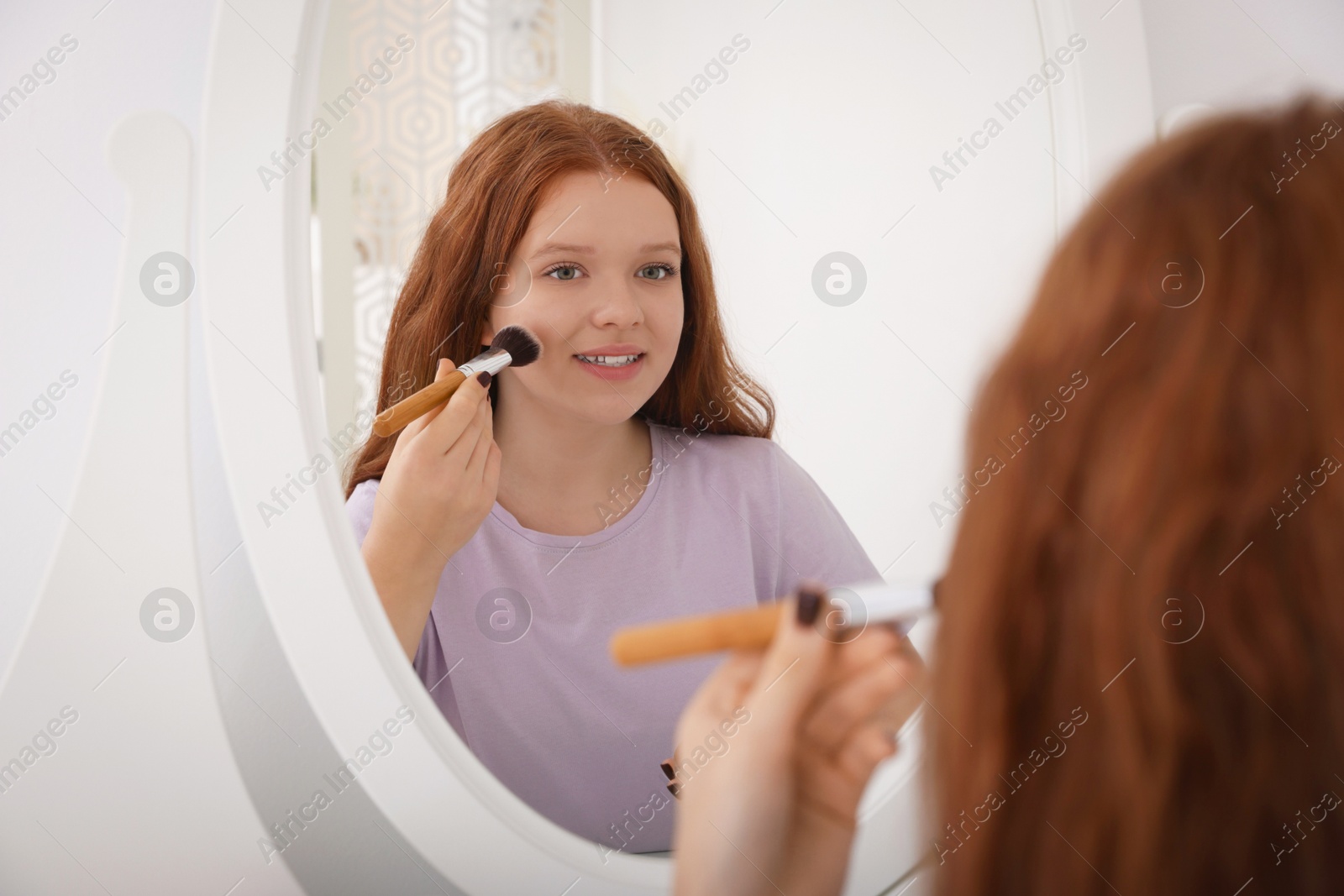 Photo of Teenage girl applying blush with brush near mirror indoors