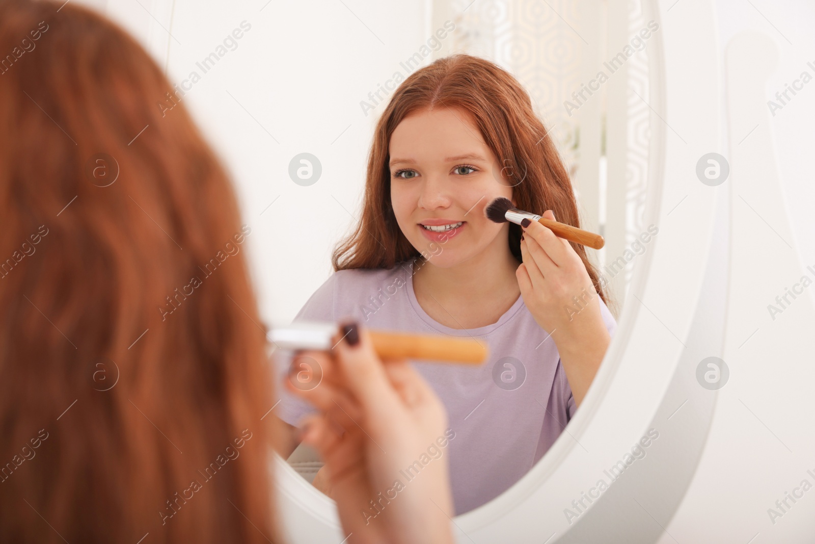 Photo of Teenage girl applying blush with brush near mirror indoors