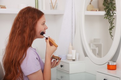 Teenage girl applying blush with brush near mirror indoors