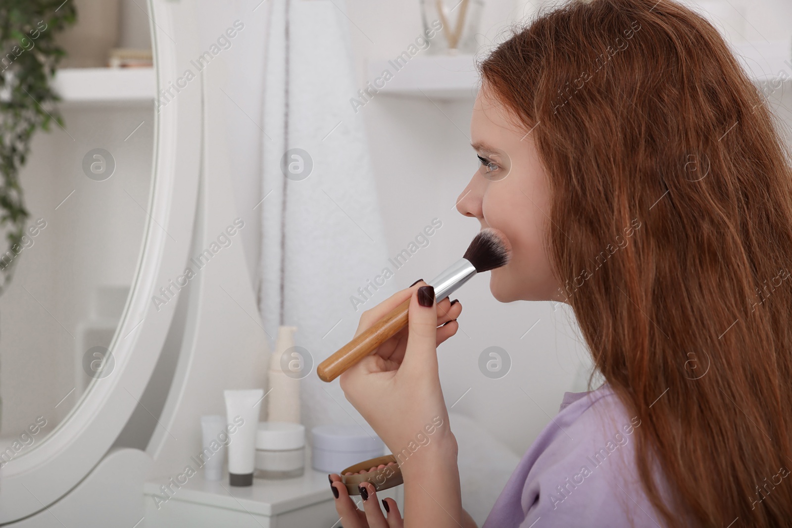 Photo of Teenage girl applying blush with brush near mirror indoors