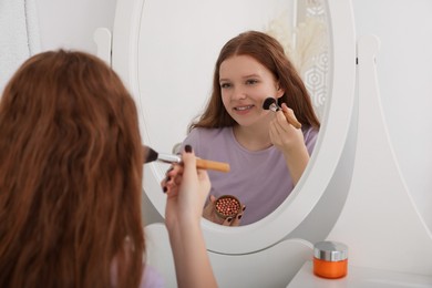 Teenage girl applying blush with brush near mirror indoors