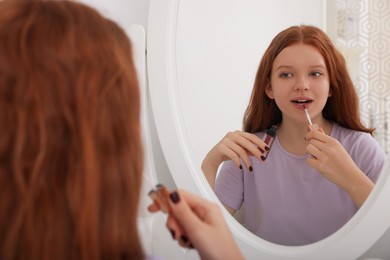 Photo of Teenage girl applying lipgloss near mirror indoors