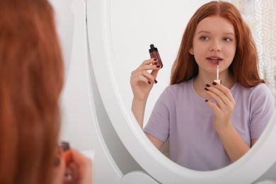 Photo of Teenage girl applying lipgloss near mirror indoors