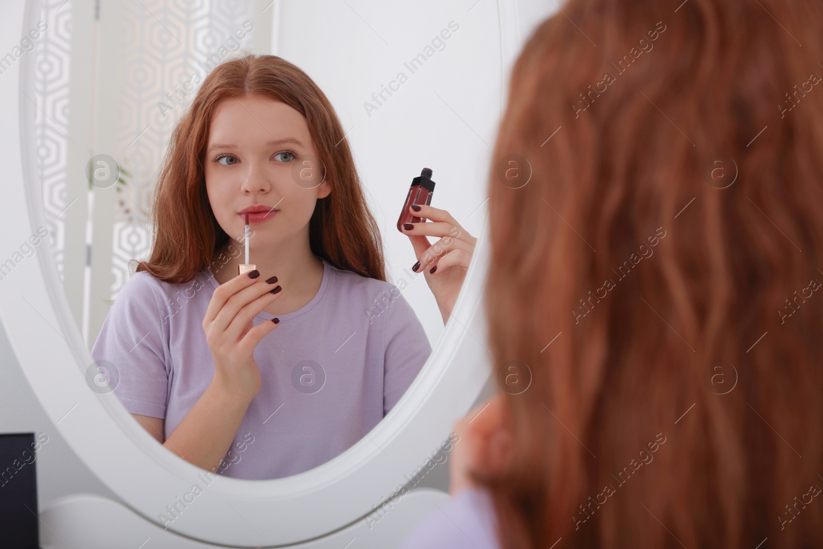 Photo of Teenage girl applying lipgloss near mirror indoors