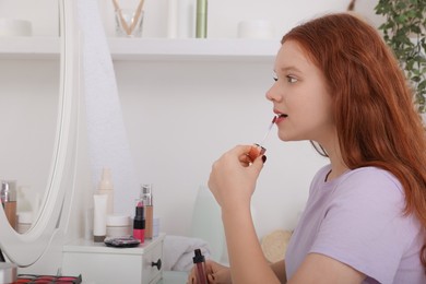 Photo of Teenage girl applying lipgloss near mirror indoors