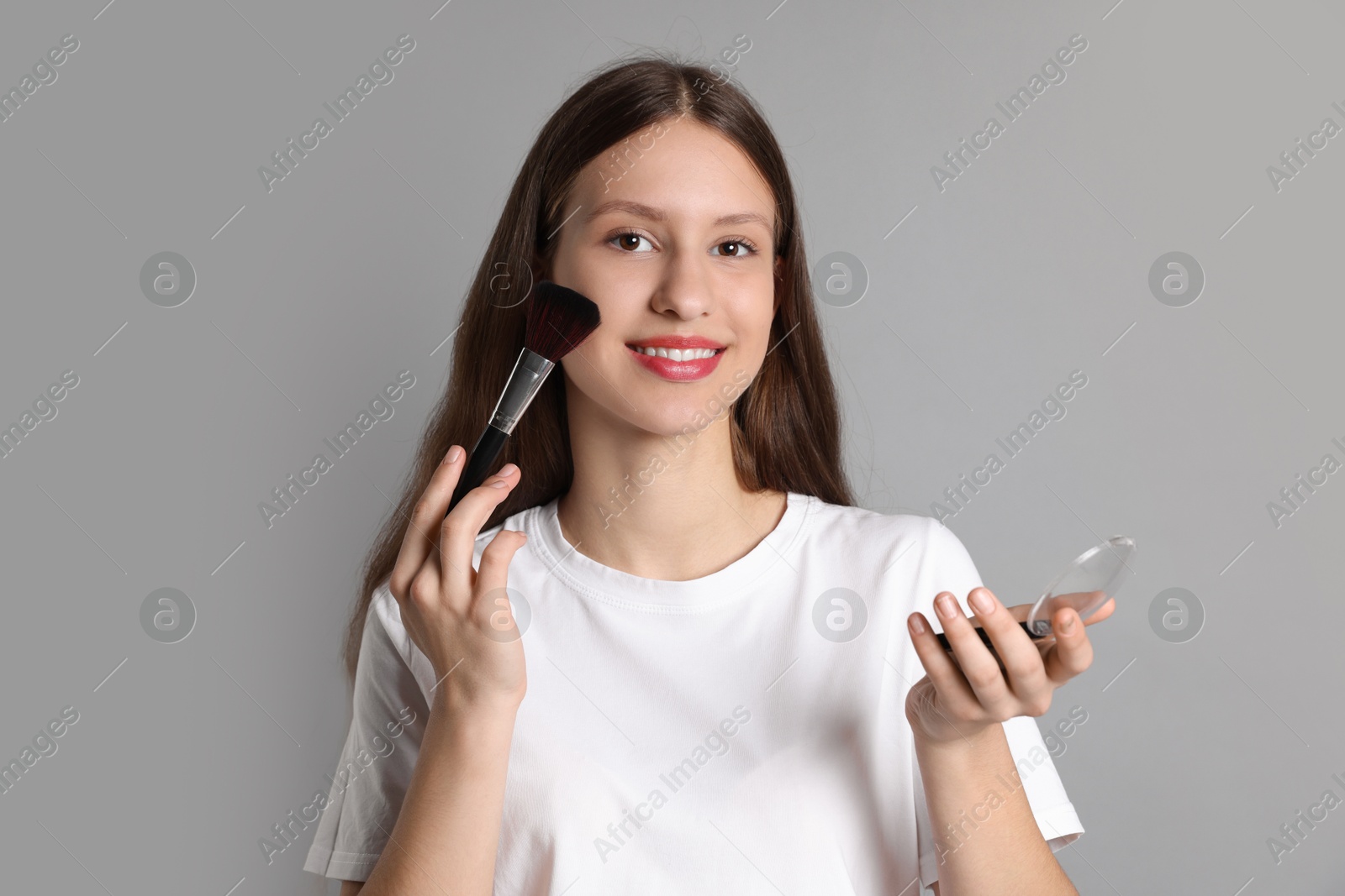 Photo of Smiling teenage girl applying blusher with makeup brush on grey background