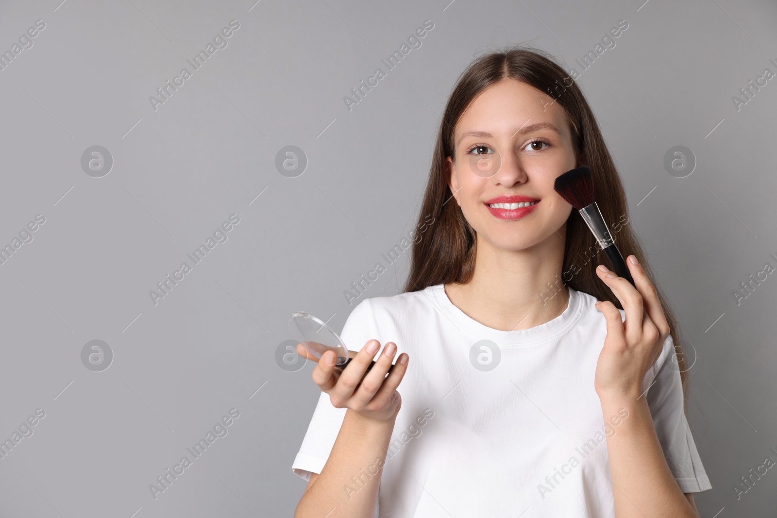 Photo of Smiling teenage girl applying blusher with makeup brush on grey background. Space for text