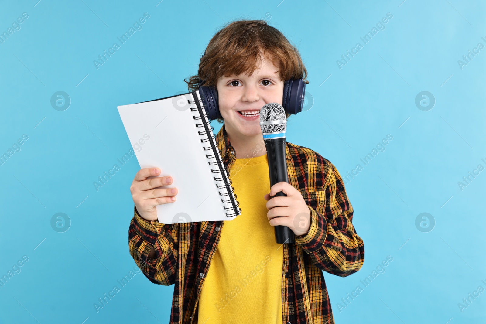 Photo of Little boy with microphone, headphones and notebook on light blue background