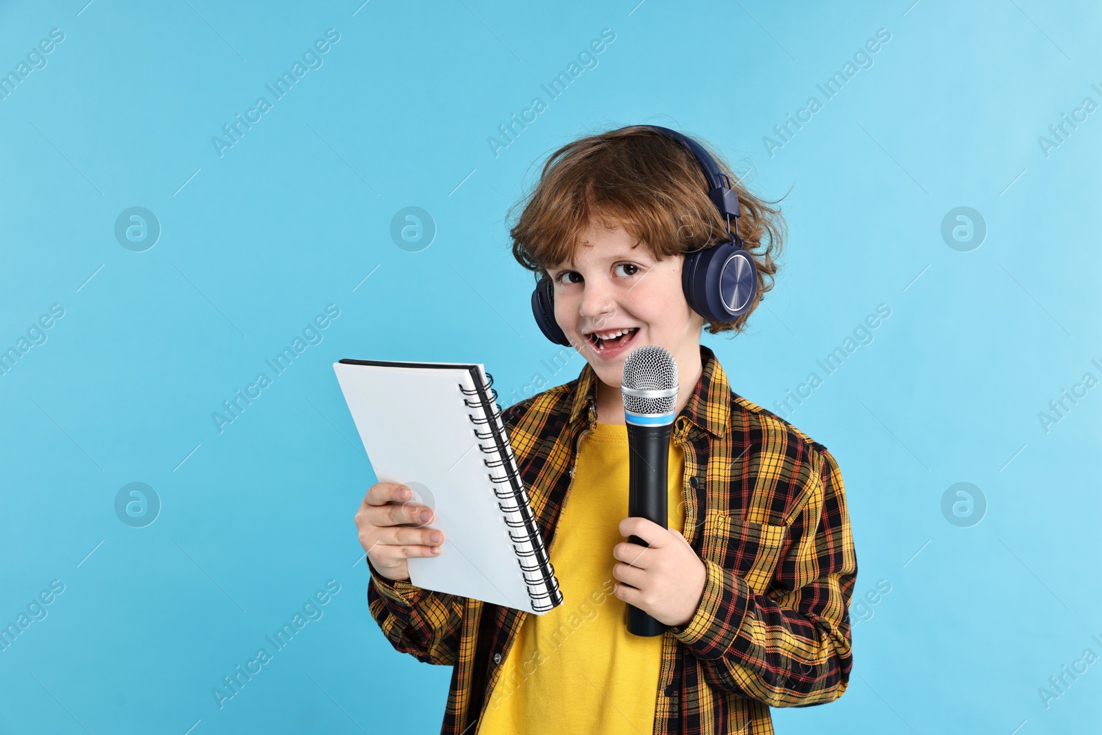 Photo of Little boy with microphone, headphones and notebook on light blue background