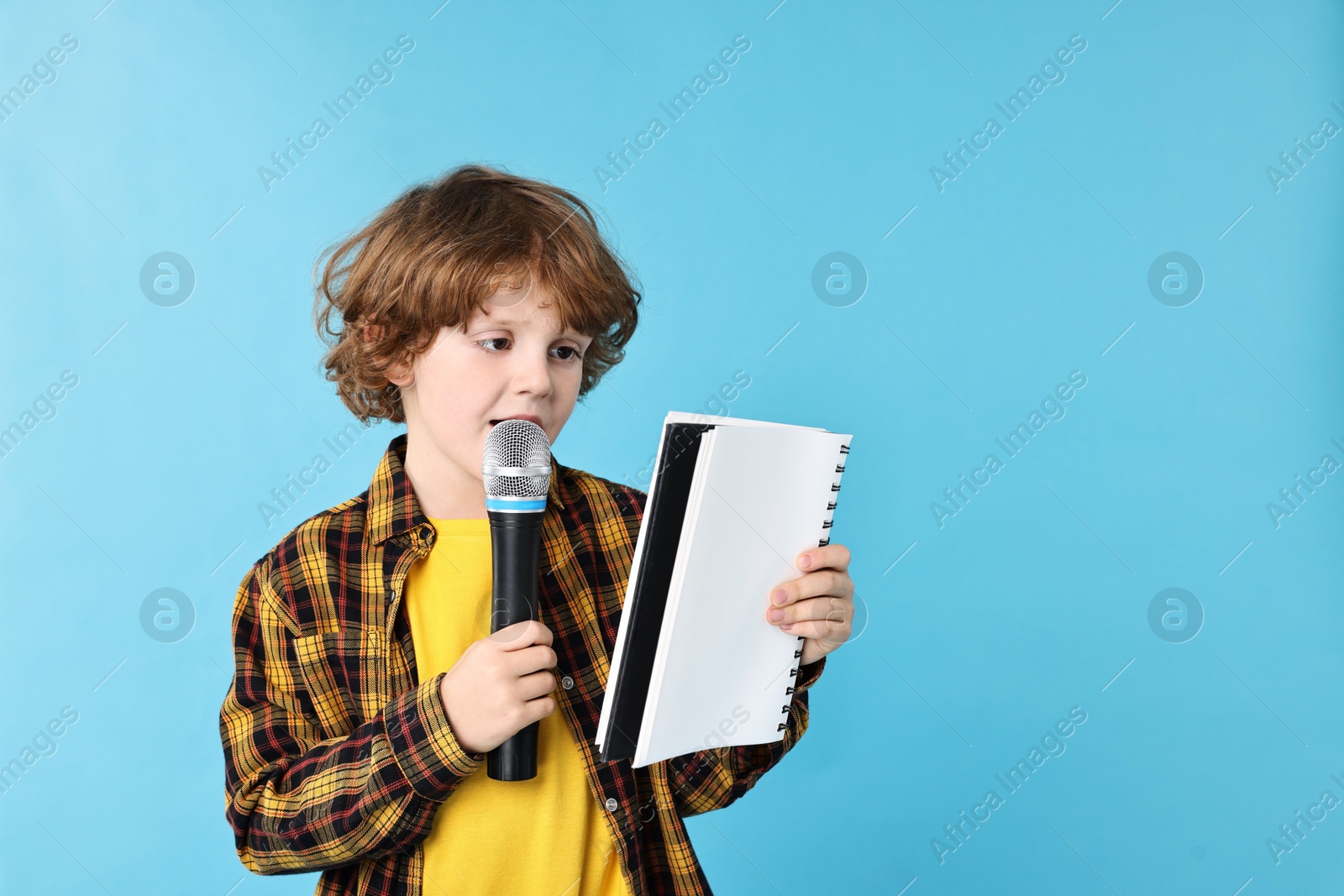 Photo of Little boy with microphone and notebook on light blue background, space for text