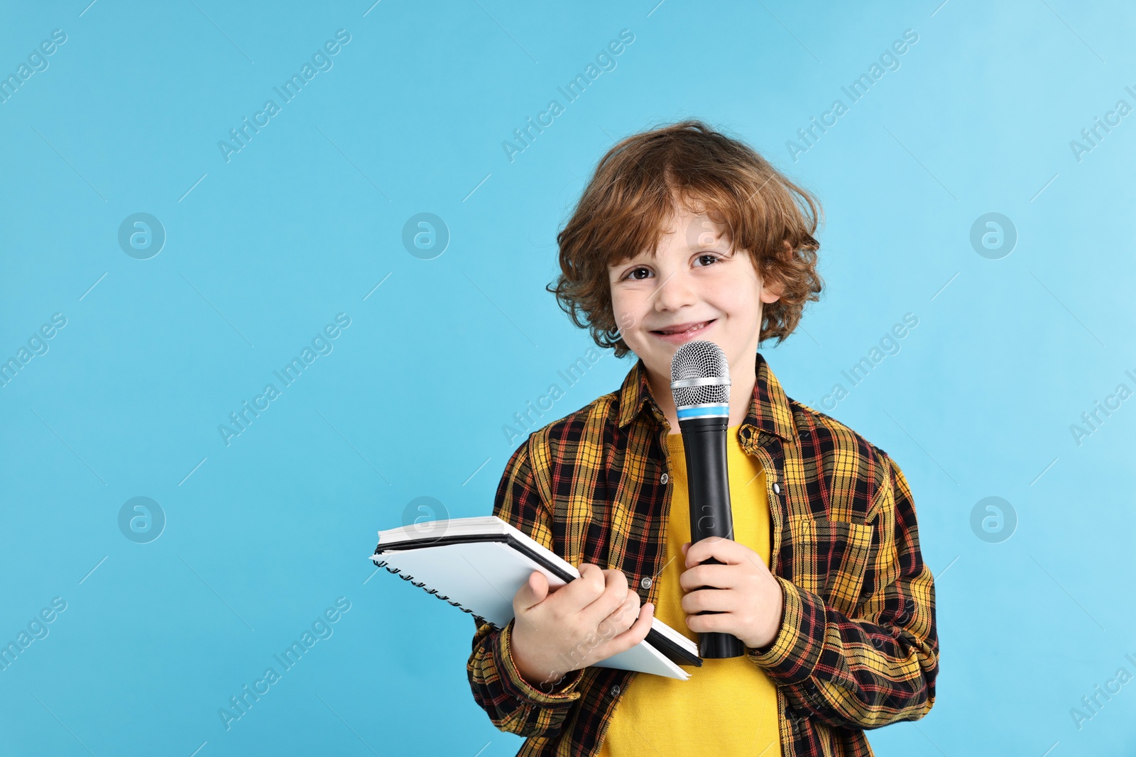 Photo of Little boy with microphone and notebook on light blue background, space for text