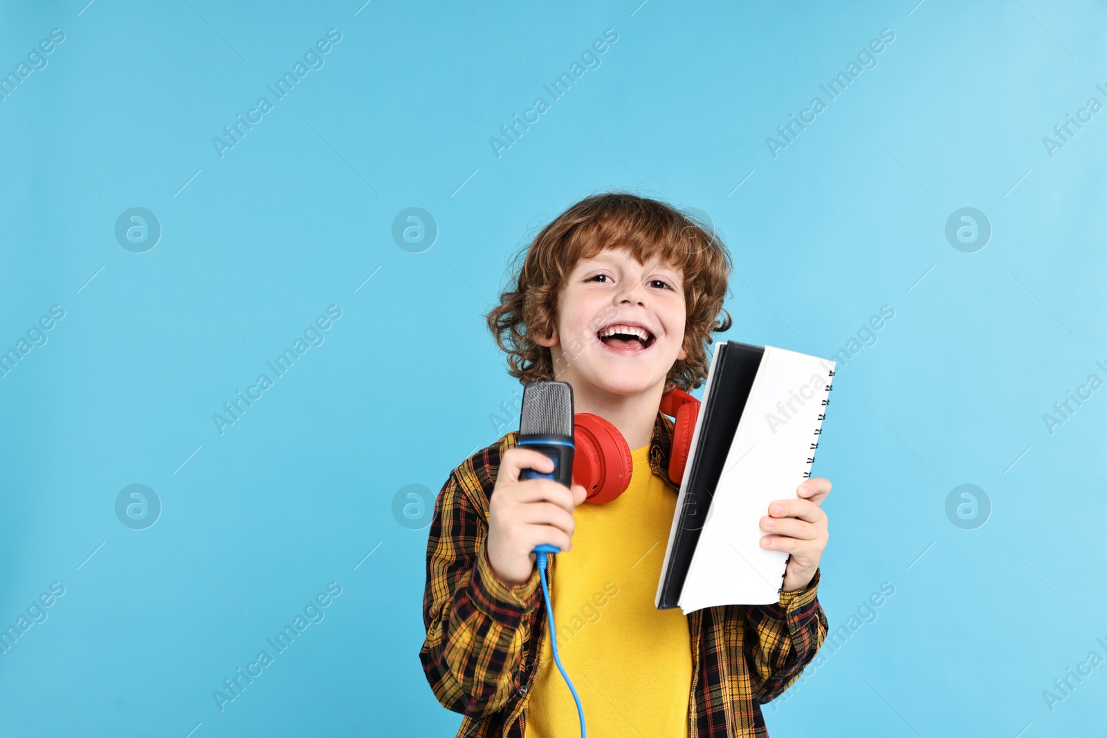 Photo of Little boy with microphone, headphones and notebook singing on light blue background